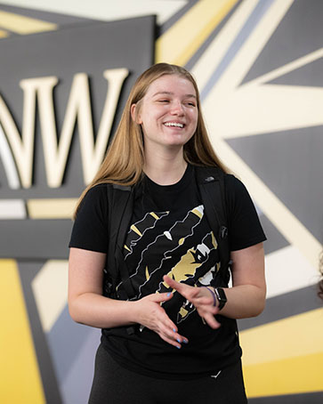 A student stands in front of the PNW mural. They are smiling and holding their hands in front of them