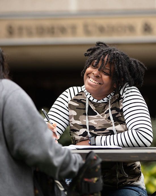 A student sits at an outdoor picnic table and is smiling at the person sitting across from them