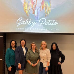 Gisele Casanova, Maureen Bauer, Nicole Schmidt, Tara Petito and Nicky Ali Jackson pose together under a projector screen that displays the logo for the Gabby Petito Foundation.