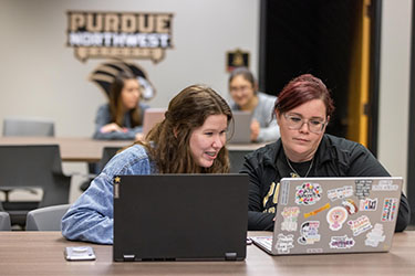 Two students sit next to each other at a table. They both have open laptops sitting on the table but are both looking at the laptop on the right.