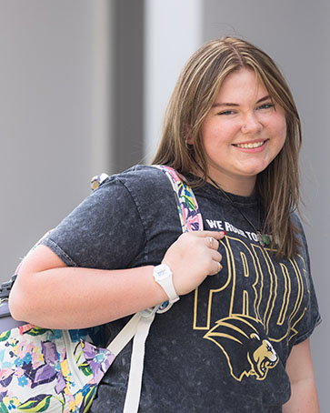 A student in a PNW Pride shirt and a floral backpack poses and smiles. They are holding onto the strap of their backpack