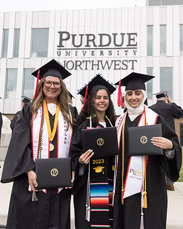 Three students pose together during commencement. They are all wearing black commencement regalia, holding their degree holders and each have a variety of tassels and stoles.