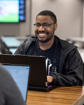 A student sits smiling at a table. Their hands are resting on a laptop.