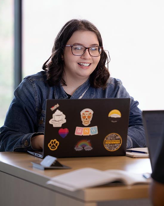 A PNW student works behind a laptop covered in students.