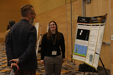Two people stand near a printed research poster. They are looking at each other while talking.