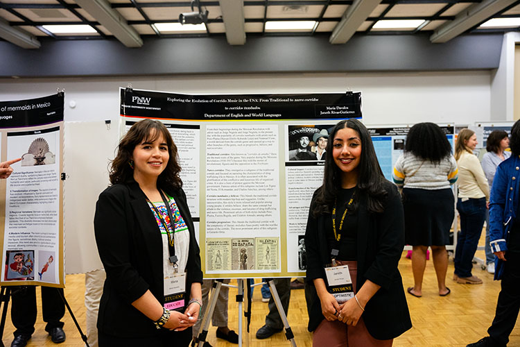 Two students stand in front of a research poster at an academic event. They are smiling and wearing conference badges. Other attendees and posters are visible in the background.