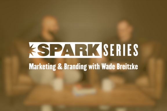 Gold text overlay over two people sitting on a couch with the text: Spark Series, Marketing & Branding with Wade Breitzke