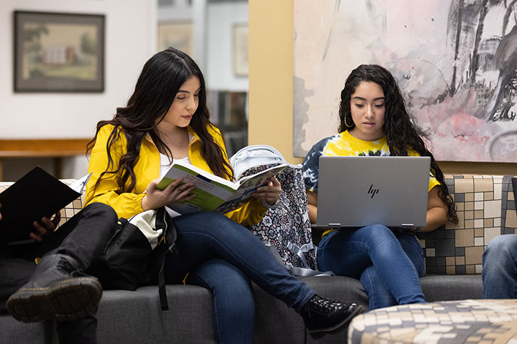 Two students sit on a couch and study. One student is holding a book and the other one is typing on a laptop.