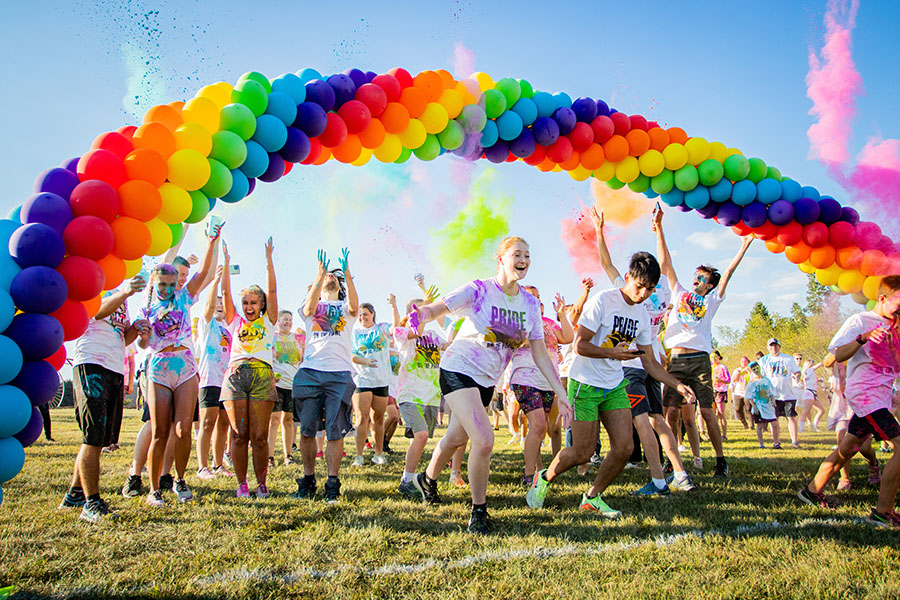 students throw color powder in the air while running under a rainbow balloon arch