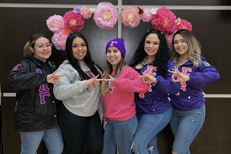 Five Sigma Lambda Gamma sisters stand together in sorority sweatshirts. They are all posing with the Sigma Lambda Gamma hand sign.