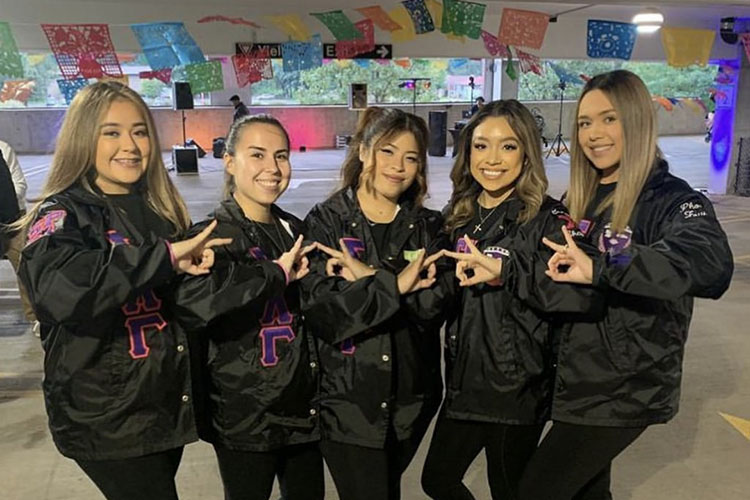 Five Sigma Lambda Gamma sisters stand together in matching black jackets with purple and pink details and black pants. They are all posing with the Sigma Lambda Gamma hand sign.