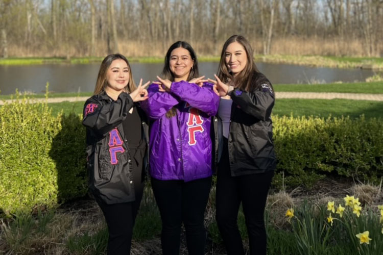 Three Sigma Lambda Gamma sisters stand together. The outer two sisters are in matching black jackets with purple and pink details and black pants. The middle sister is in a purple jacket with pink accents and black leggings. They are all posing with the Sigma Lambda Gamma hand sign.