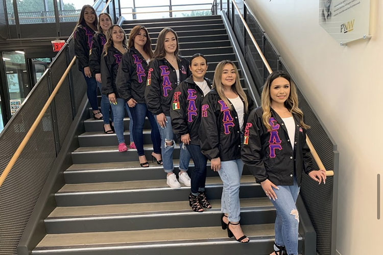 8 Sigma Lambda Gamma sisters pose on a stair case. They are standing diagonally across the staircase so everyone is seen. They are all wearing jeans and matching black jackets with pink and purple details.