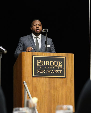 Man in a suit stands at a podium. The front of the podium reads "Purdue University Northwest"