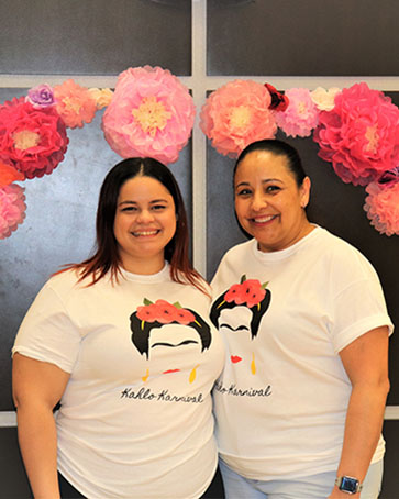 Two women pose under a balloon arch in matching Kahlo Karnival shirts