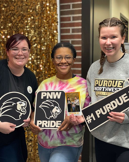 Three students pose together holding PNW photo props. They are in front of a gold background