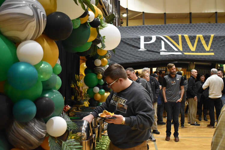 A person picks up items from a table. There is a green, black, white and gold balloon arch above the table