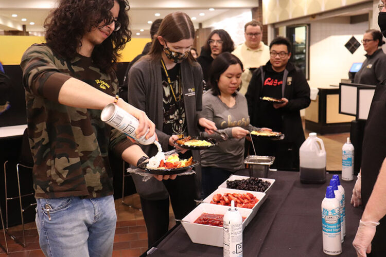 A student sprays whipped cream onto a plate of pancakes. There is a line of other students in the background