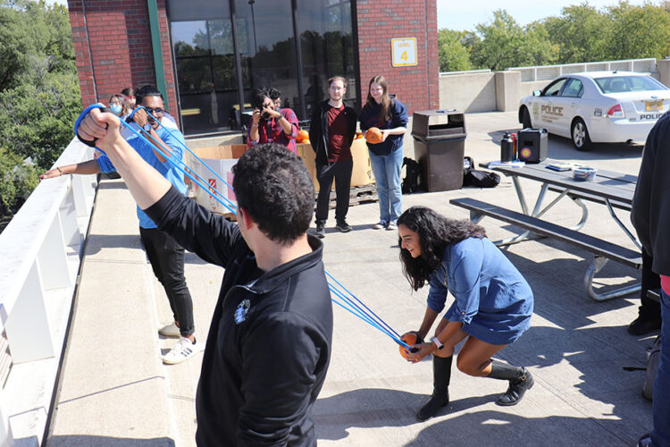 Two people hold the strings of a catapult while a student pulls the center back with a pumpkin to launch it