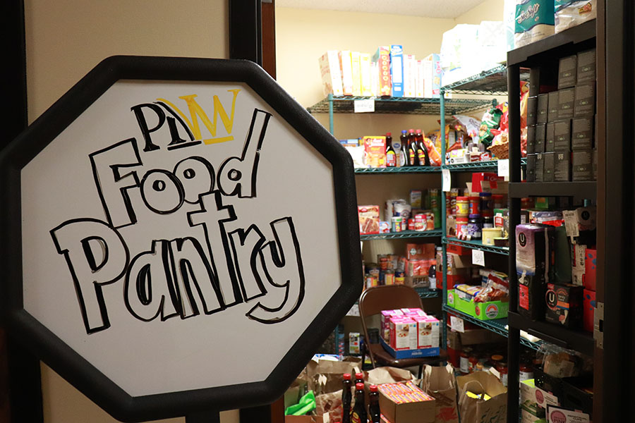 An octagon shaped white board with "PNW Food Pantry" written on it. The food pantry is in the background with fully stocked shelves.