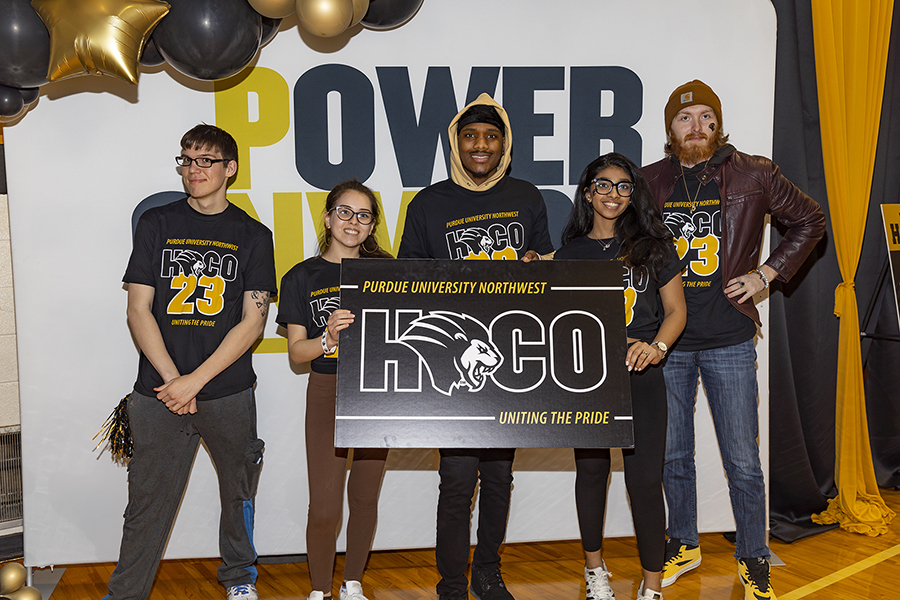 Five students stand in front of a PNW "Power Onward" back drop. They are wearing matching homecoming t-shirts and are holding a black homecoming sign