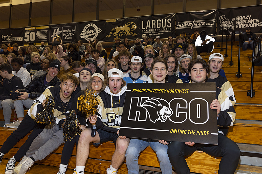 Students sitting in bleachers. The students in the front row are holding a black homecoming sign.
