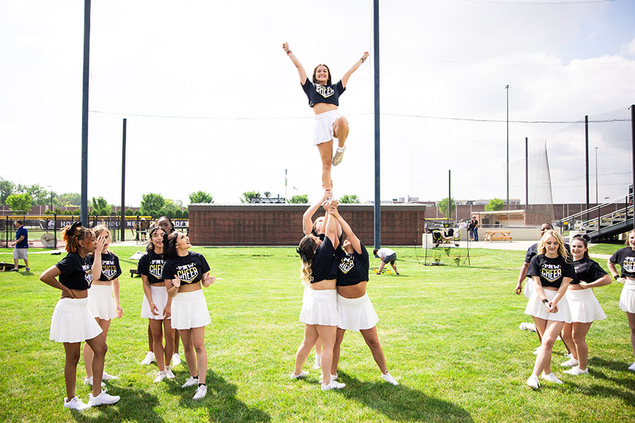 A PNW cheerleader stands and smiles while being lifted into a pyramid. Other cheerleaders smile and watch.