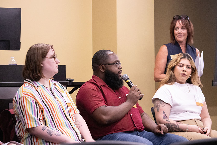 Three alumni sit in a chairs. A staff member is standing off to the right side