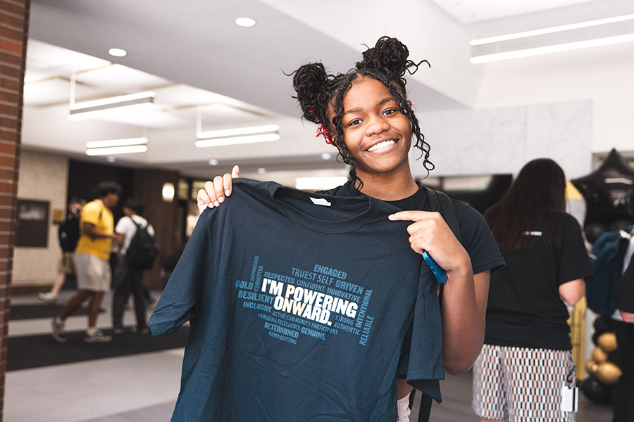 A student smiles and holds up an "I'm Powering Onward" t-shirt