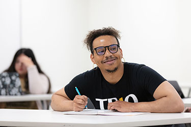 A student in a black TRIO t-shirt sits and leans on a table. They are holding a blue pencil and there is paper on the table.