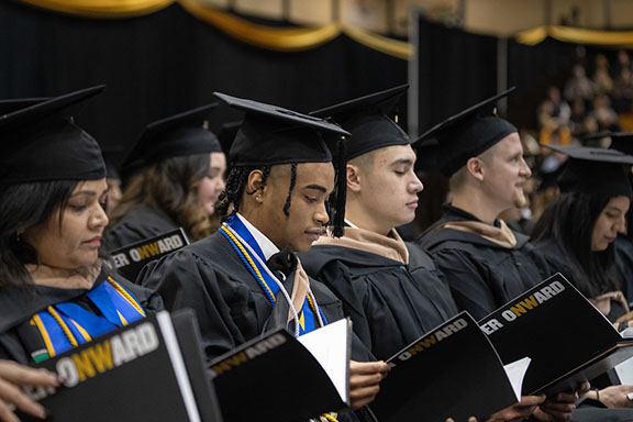 Students sit in rows during commencement. The photo focuses on two students who are looking at the commencement booklet they are holding. Everyone is wearing black commencement regalia.