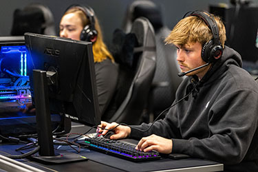 Two students sit at esports computers. They both have headphones on.