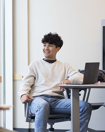 A student in a white sweater and light blue jeans sits in a computer chair and looks out of a window.