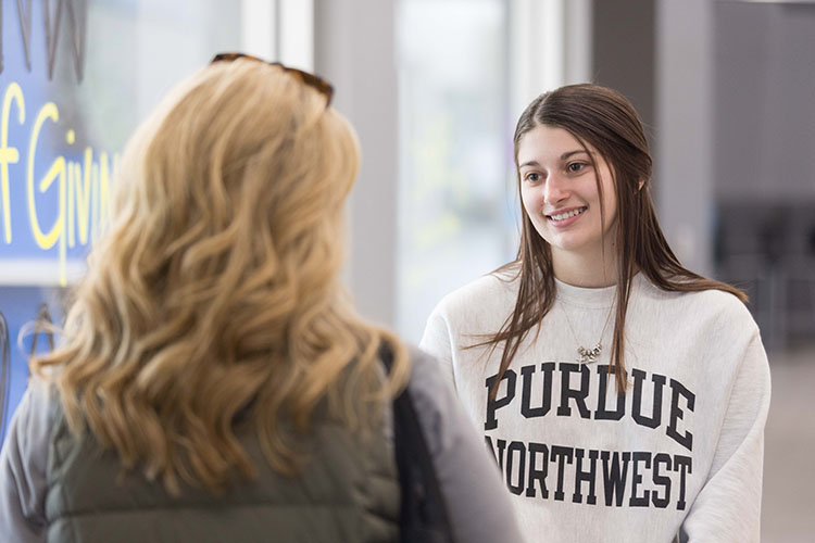 Two students stand and talk. The student facing the camera has long black hair and is wearing a Purdue Northwest sweatshirt. The student facing away from the camera has long blonde hair and is wearing a gray long sleeve shirt.