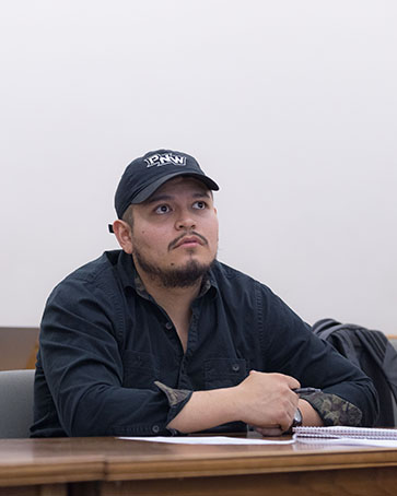 A student in a black button down shirt and a black PNW hat sits in class.