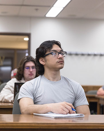 A student in a gray t-shirt and glasses sits at a desk in a classroom.