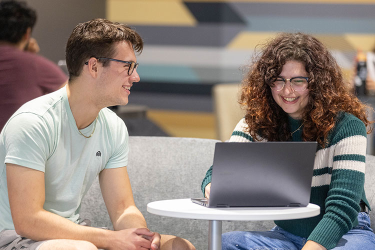 Two students sit together at a table and work on a laptop