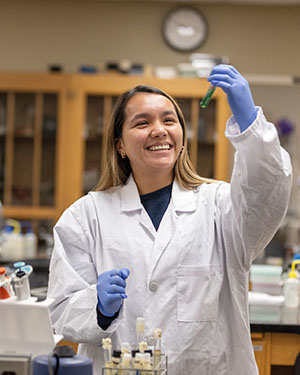 A PNW student in a lab coat looks up at a test tube.