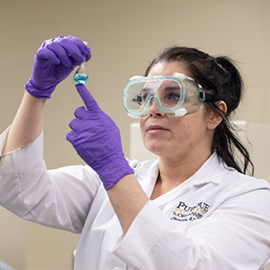 A PNW student in a lab coat and protective goggles holds up a tube of liquid.