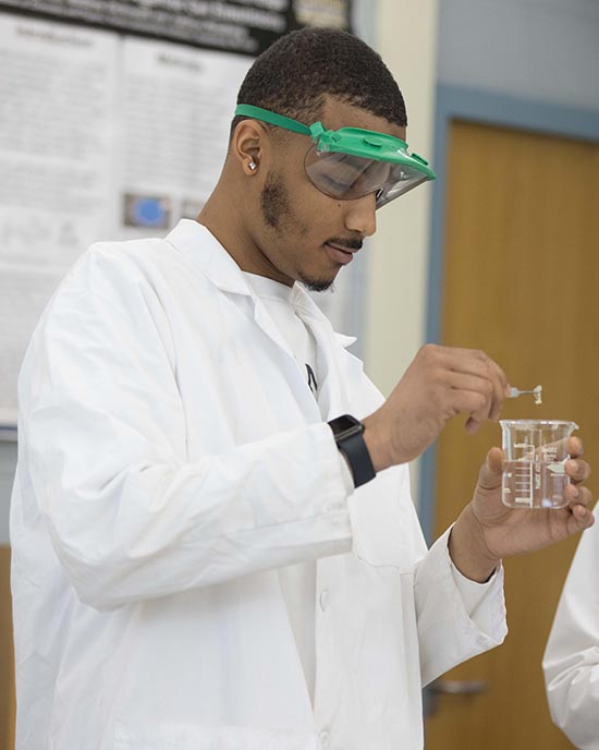 A student in a lab coat inspects a flask and adds mixture inside.
