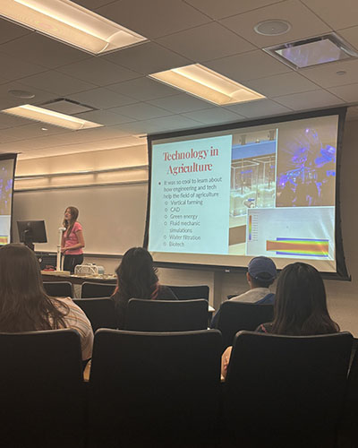 Students sit in a lecture style hall and listen to another student who is giving a presentation.