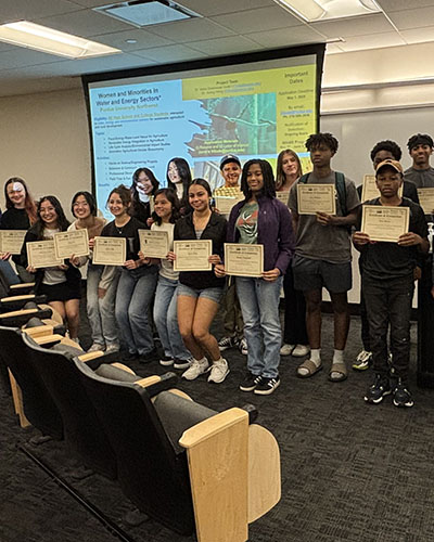 Students stand in front of a projection screen. They are all posing together and holding up completion certificates.