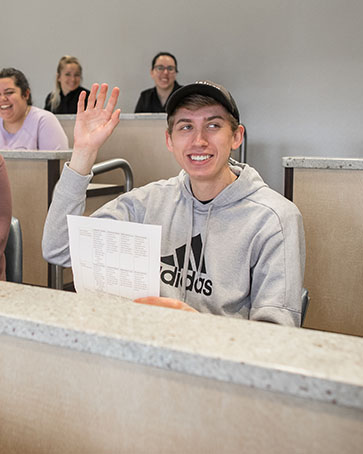 A student sits in class. They are raising their hand and holding a sheet of paper.