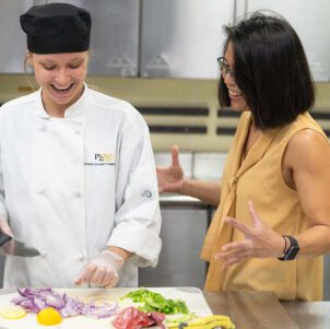 A student stands with an HTM instructor in the kitchen. The student is chopping food on a chopping board.