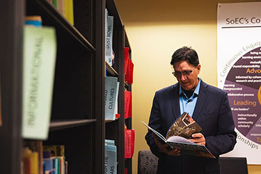 David Pratt stands next to a bookshelf. He is holding a book and looking at it.