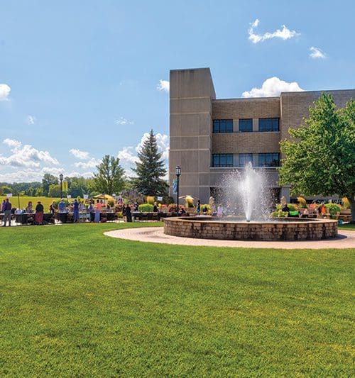 People gather in front of a building and fountain on PNW's Westville campus on a sunny day.