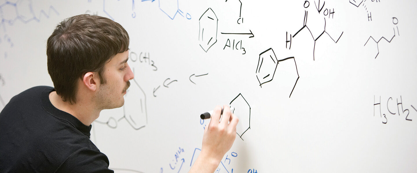 A student writes a chemistry equation on a white board.