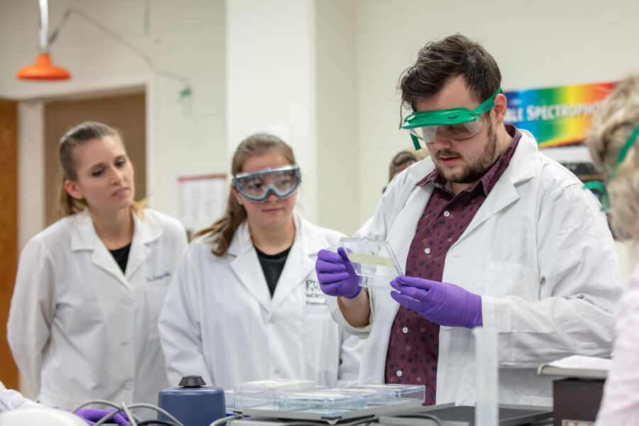 Students in white lab coats and goggles work in a lab