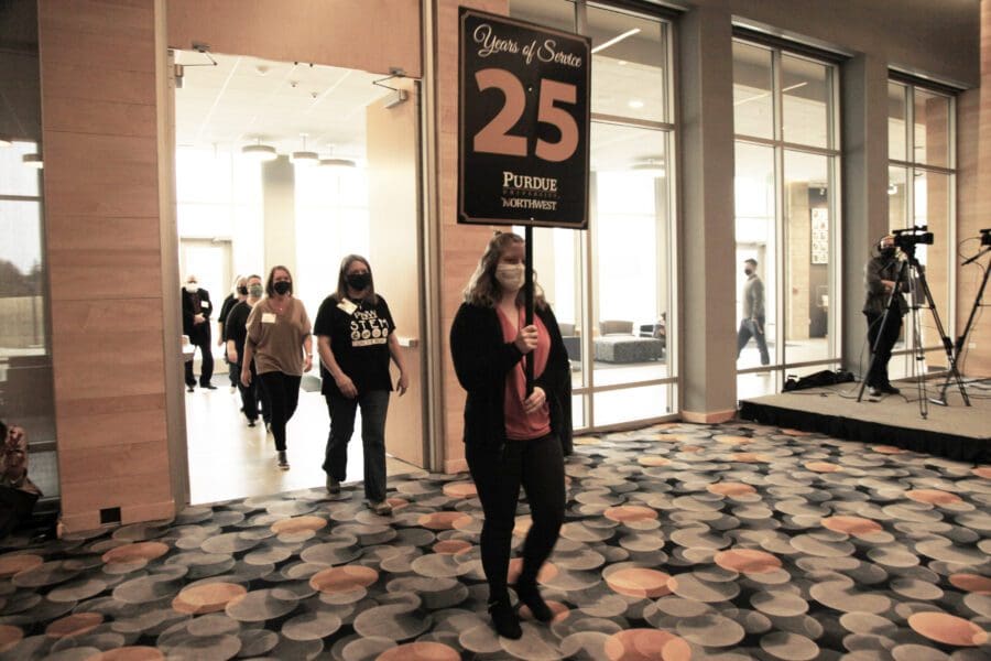 Three individuals marching through a room. The first person is holding a flag stating "25 Years of Service"