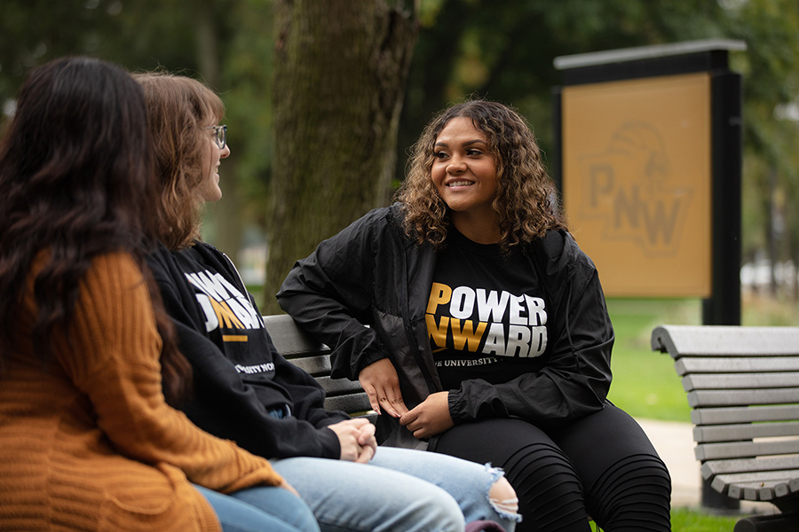 PNW students talk together on a bench. One is wearing a "Power Onward" shirt.